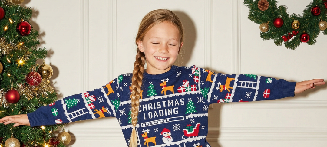 Smiling girl with a braid wearing a navy Christmas Loading jumper, arms outstretched beside a decorated tree and wreath