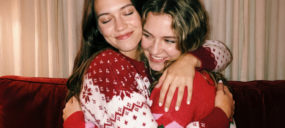 Two smiling women in festive red and white Christmas sweaters hugging on a couch